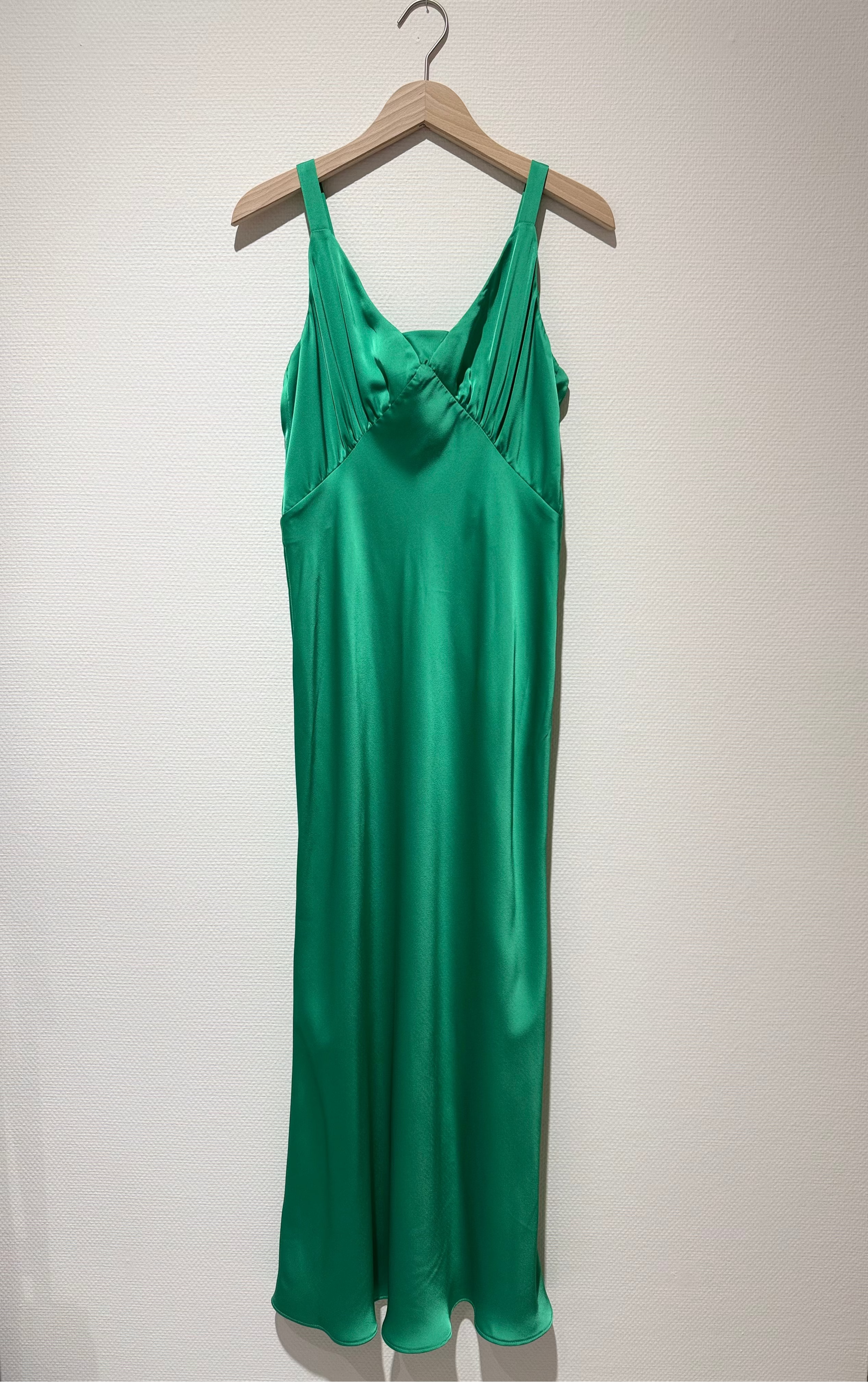 A vibrant emerald green satin slip dress hanging on a wooden hanger against a plain off-white wall.