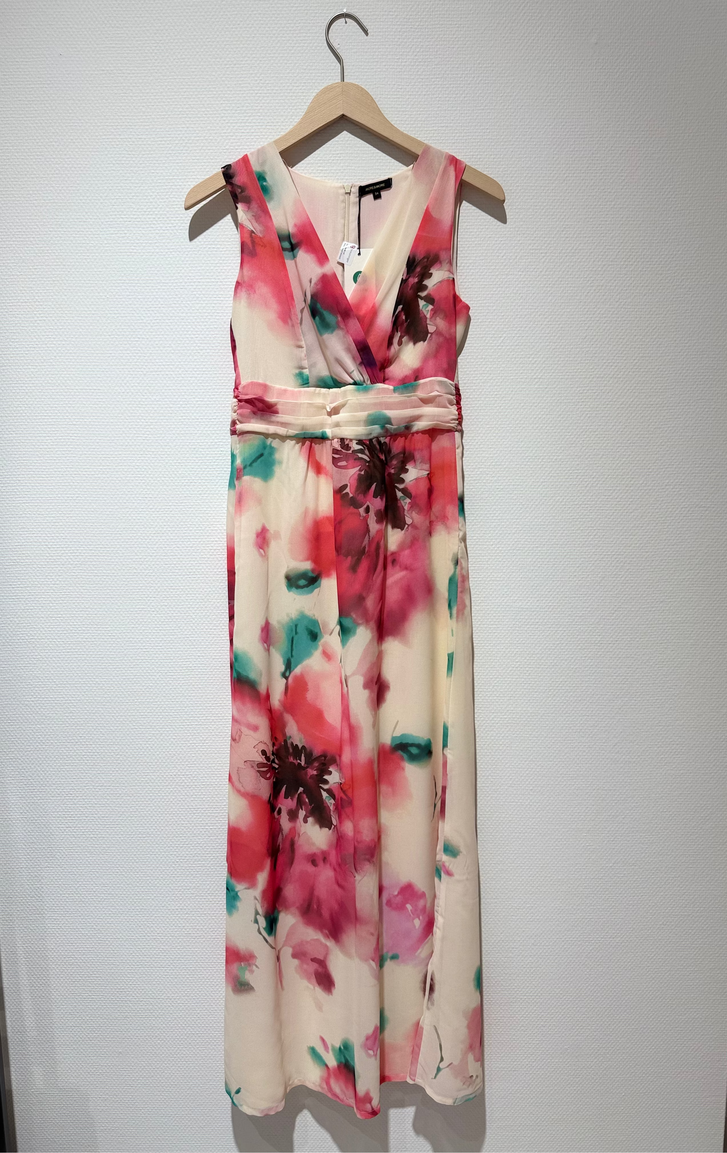 A sleeveless V-neck maxi dress with a watercolor floral print in pink, red, and teal on a cream base, hanging on a wooden hanger against a white wall.