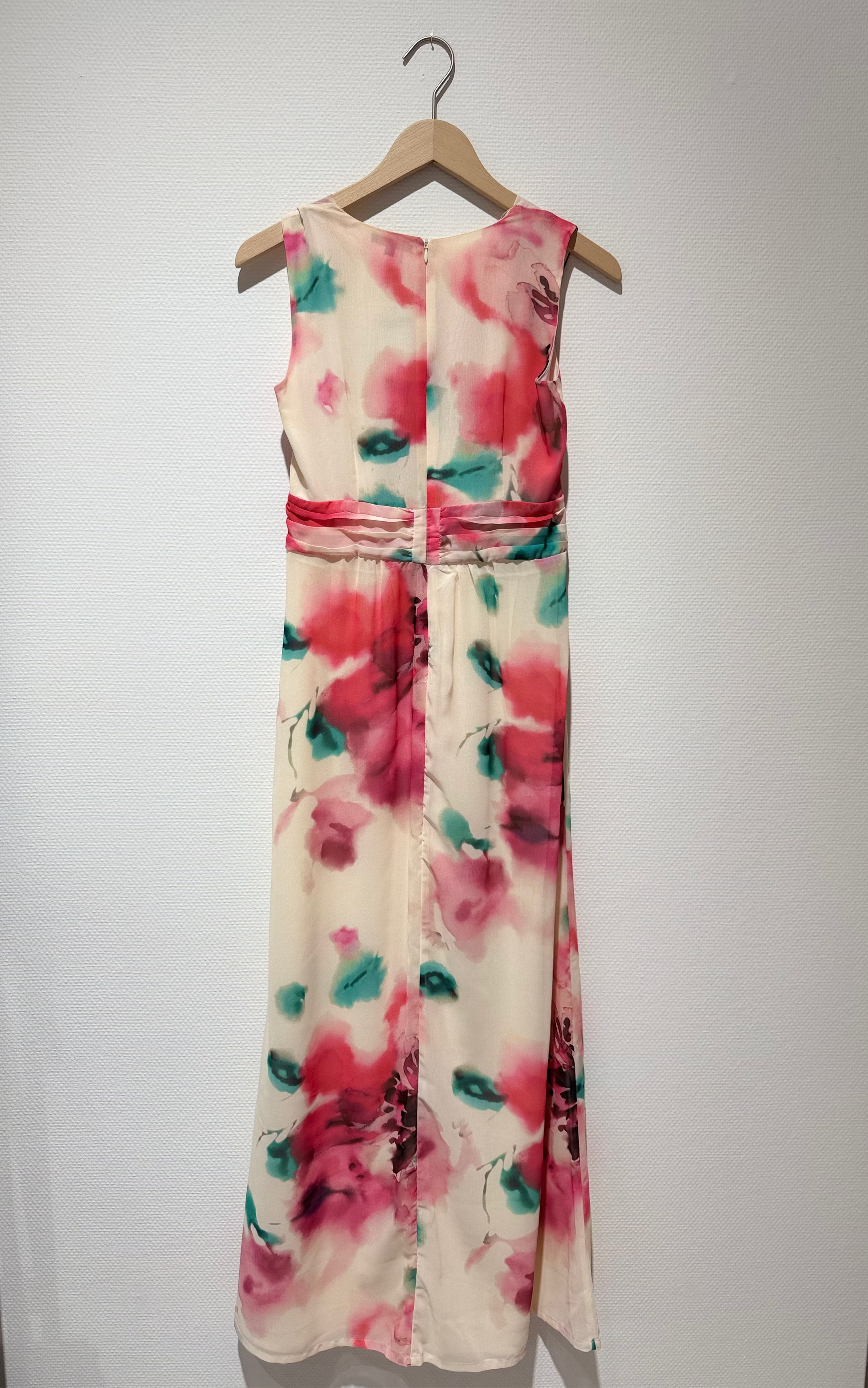 A sleeveless maxi dress with a pink and teal watercolor floral print on a cream background, hanging on a wooden hanger against a white wall.