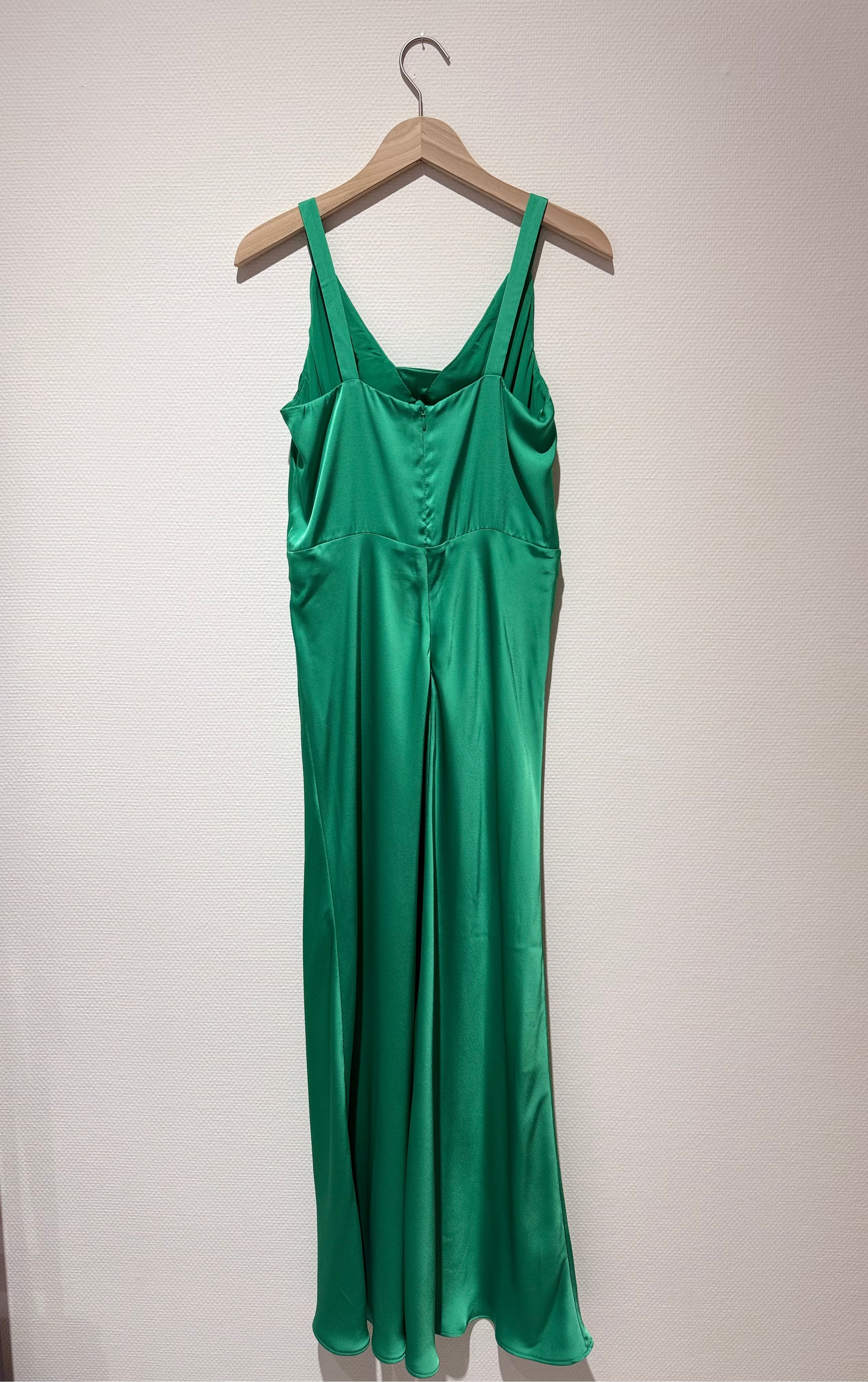 A green satin slip dress hanging on a wooden hanger against a plain off-white wall.