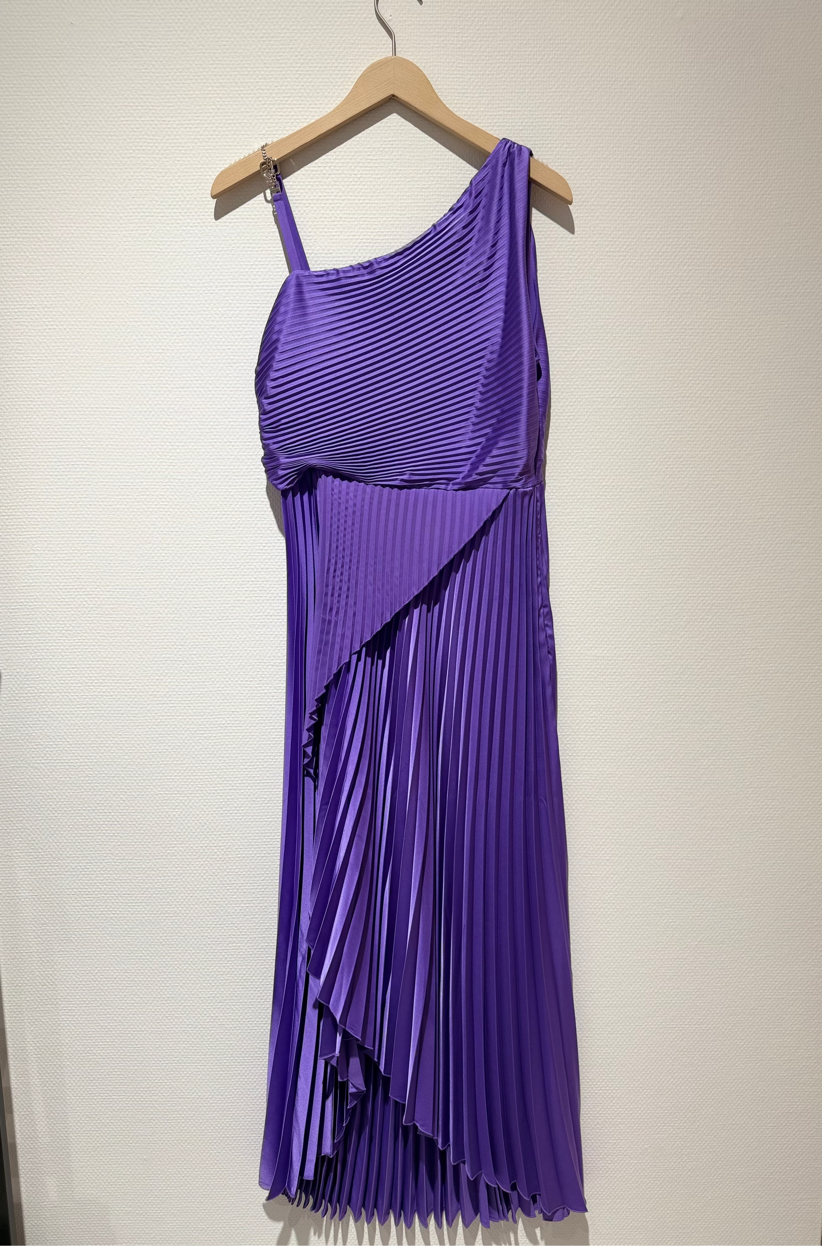 A purple pleated one-shoulder midi dress hanging on a wooden hanger against a plain off-white wall.