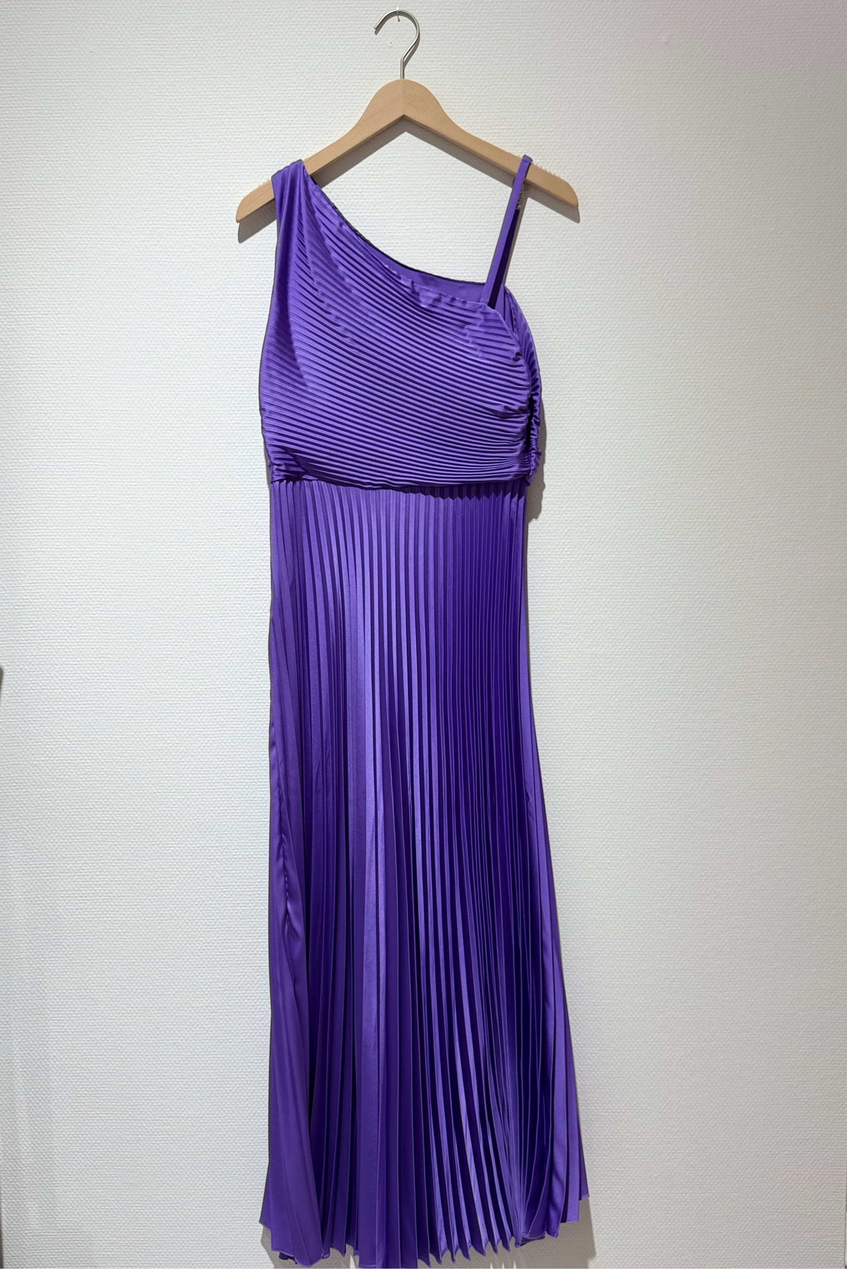 A purple pleated one-shoulder maxi dress hanging on a wooden hanger against a plain white wall.