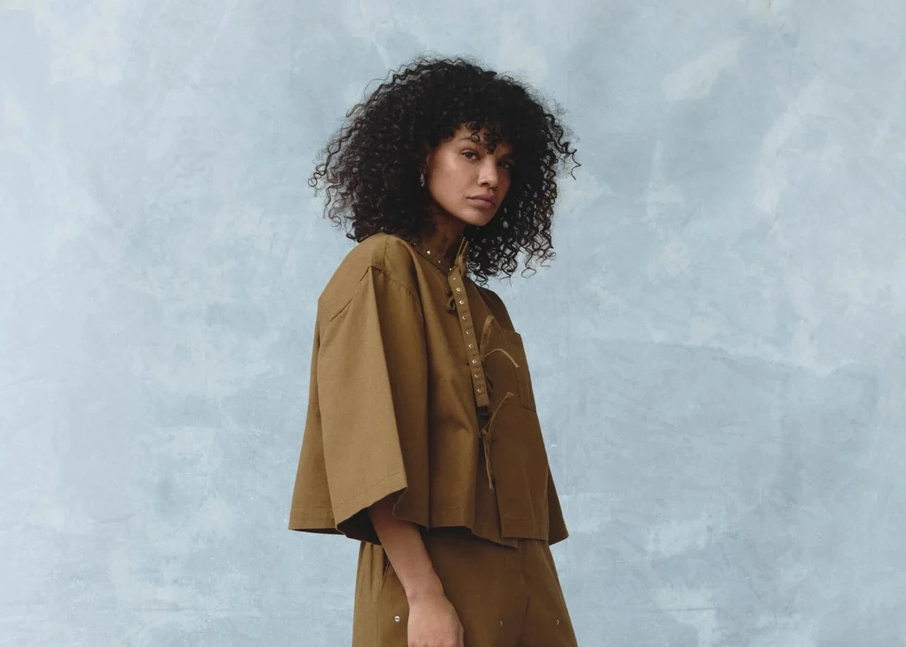 A model with curly dark hair stands in profile wearing a matching brown two-piece outfit consisting of a cropped short-sleeve top and wide-leg trousers with small white embroidered dots, set against a textured light blue studio backdrop.